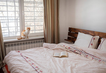 Interior of bedroom. Light bedding, book on bed and cat on window