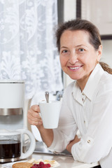 Mature woman   eating coffee with cookies in   kitchen.
