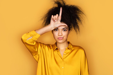 Girl making fun of people with fingers on forehead doing loser gesture mocking and insulting. Photo of african american girl on yellow background