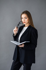 Young pretty girl in business style clothes stands on a gray background with a notebook and a pen in her hands. Business style