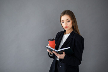 Beautiful young woman in business clothes is holding a cup of coffee, looking at camera and smiling, standing against gray wall