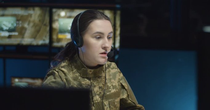Caucasian military woman in headset sitting at computer screen and talking via videochat in controlling center of army. Soldier speaking via webcam in military office. Female officer in dispatch room.