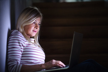 Senior woman with laptop indoors at home in the evening, working.