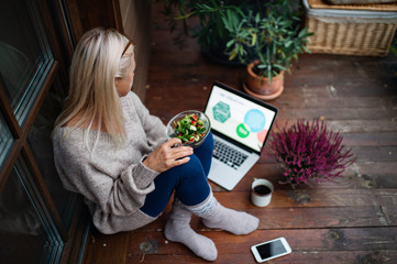 Senior woman with laptop sitting outdoors on terrace, working.