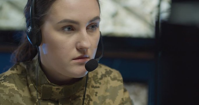 Portrait of Caucasian young woman in headset sitting at computer screen in monitoring room in army and then looking at camera. Close up of female soldier despatcher in military center.