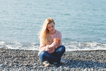 outdoor portrait of pretty blonde young woman in pink knitted sweater sitting at sea shore alone and drinking coffee or tea