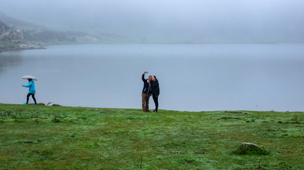 Pareja haciéndose una auto foto el los lagos de Covadonga mientras pasa una chica con paraguas en...