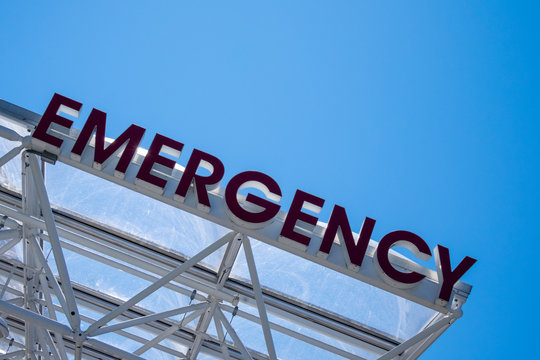 A large red emergency glass sign against a bright blue sky. The exterior hospital emergency department sign hangs on the top of a glass roof covering. The red letters have lights inside them. 