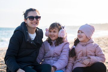 a young woman and two little girls in jackets and earmuffs play and hugs on the beach