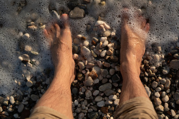 Sea foam washes men's feet standing on a pebble beach
