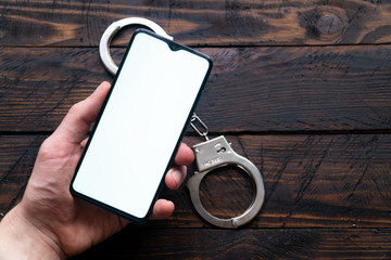 Man's hand holds a modern smartphone with a white screen, handcuffs lie against a wooden background