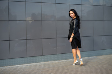 Street fashion spring look. Beautiful young woman with long hair wearing elegant black dress and fashionable black fringed leather jacket posing on urban wall background.