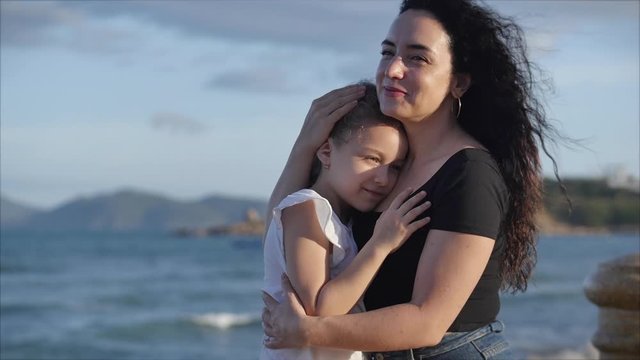 Happy family, mother and a cute daughter in their gentle embrace sit on a bench near the sea, the child with his beloved mother smiles and hugs each other tightly. Concept love of parents and children