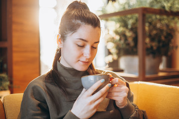 portrait of a beautiful woman drinking coffee or tea with a large mug sitting in a room where a lot of bright sunlight and sunlight go through the windows and create a warm light atmosphere