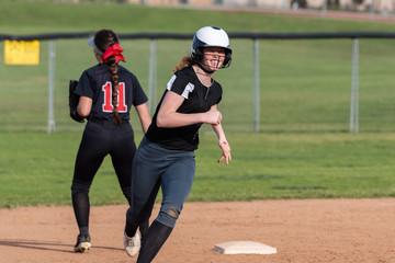 High school female softball player runs at full speed around second base with smiling expression on face.