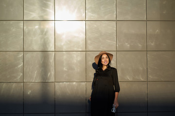  Young attractive cheerful girl with charming smile wearing youth black dress in stylish hat. Model in trendy outfit posing on urban wall background and looking at camera. Outdoor portrait.