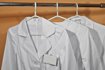 White medical gowns hang on hangers. Clothes on orange background. Bathrobes with an empty badge.