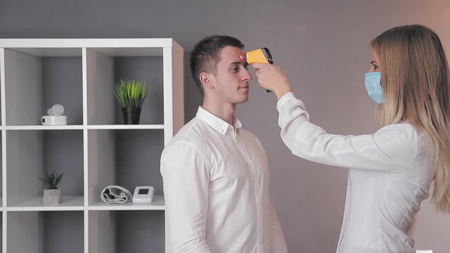 Doctor in a white coat and a medical mask measures the temperature of a patient in the medical office with an infrared non-contact thermometer.