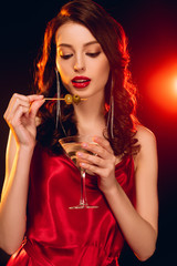 Beautiful woman holding stick with olives and glass of martini on black background with lighting
