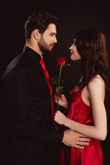 Side view of handsome man hugging elegance girlfriend with rose isolated on black