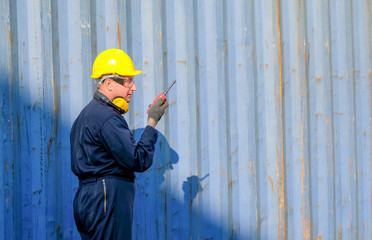 Technician or engineer with blue uniform and yellow helmet use walkie-talkie to contact with co-worker in cargo container shipping area.