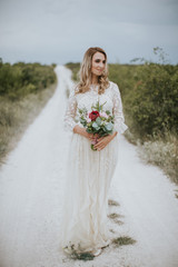 Fototapeta na wymiar Smiling young blonde caucasian bride in a white, modern lightweight dress posing. Wedding bouquet in her hands. Happy vintage hipster bride, warm colors.