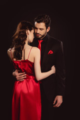 Handsome man in suit hugging by waist elegant girlfriend in red dress isolated on black