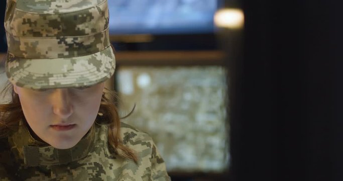 Close up of Caucasian young woman in army uniform and hat sitting in monitoring room and working at computer, then talking in walkie-talkie. Female soldier speaking in radio transmitter.