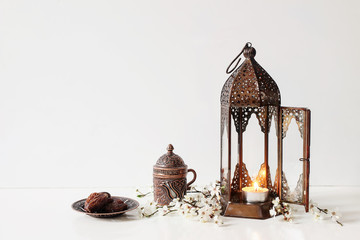 Ramadan Kareem greeting card, invitation. Bronze plate with dates fruit, white flowers, prunus tree blossoms, cup of tea and glowing Moroccan lantern on table background. Iftar dinner. Eid ul Fitr.