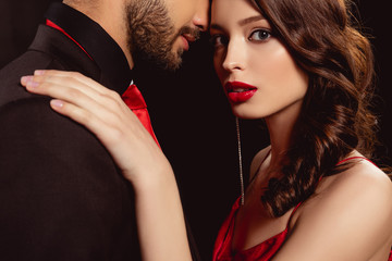 Side view of beautiful girl with red lips looking at camera while embracing boyfriend isolated on black