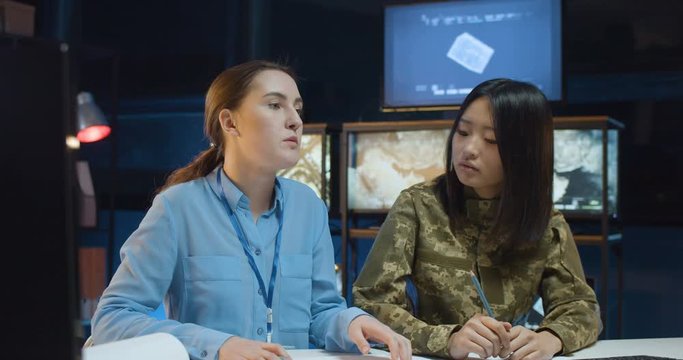Female Caucasian office worker and soldier sitting together at desk in monitoring center with screens and studying some militarian documents. Women talking and discussing army strategy.