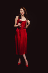 Full length of beautiful woman in dress holding glass of red wine and smiling at camera isolated on black