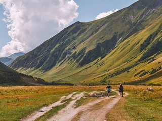 Two tourists walk down the green valley on a sunny day in Caucasus mountains