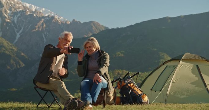 Senior cacasian couple having a rest on top of a mountain, taking a picture or having video chat on smartphone, travelling together after retirement - pension, recreational pursuit, tourism concept 4k