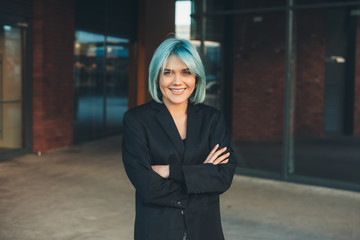 Charming caucasian businesswoman with blue hair is posing with crossed hands while looking at camera