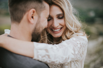 Smiling bride and groom spending time together. Posing on the mountain hills background. Dressed in white dress beautiful blonde caucasian bride and handsome groom. Hugs, kissess and enjoy the company