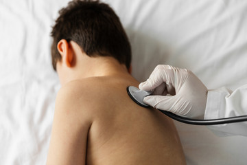 latex gloved hand of a female doctor with a phonendoscope (stethoscope) on a child's back to listen. in the hospital bed or home. home doctor. medical inspection.