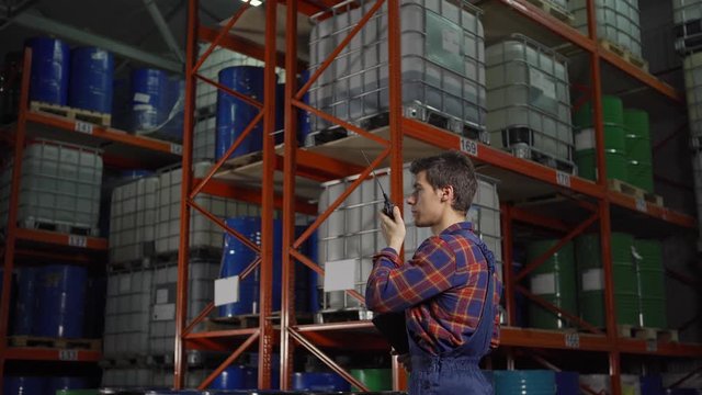 Side view tracking shot of young warehouse worker in uniform talking on walkie-talkie standing by racks with steel barrels and containers in factory storage