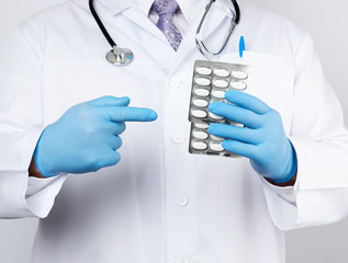 adult doctor therapist is dressed in a white uniform coat and blue sterile gloves is standing and holding a stack of pills in blister packs