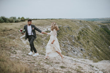 Smiling bride and groom spending time together. Posing on the mountain hills background. Dressed in white dress beautiful blonde caucasian bride and handsome groom. Hugs, kissess and enjoy the company