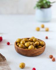 Food photo of green olives in a wooden plate on a light background, nuts and a green plant. There is a place for text.