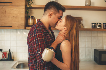 Lovely ginger woman with freckles is kissing her lover while drinking a cup of tea in the kitchen