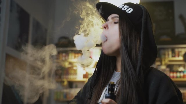 Young pretty woman in cap smoke an electronic cigarette at the vape shop. Hip-hop style. Closeup.
