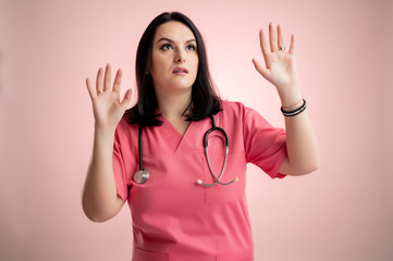 Beautiful woman doctor with stethoscope, wearing pink scrubs showing fear, with her arms up