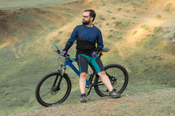 Fototapeta na wymiar Cyclist in shorts and jersey on a modern carbon hardtail bike with an air suspension fork standing on a cliff against the background of fresh green summer forest