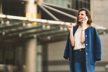 Fototapeta premium Caucasian business woman speaking by phone. Waist up portrait of a successful European business woman woman, talking on the phone, standing on glass background, modern office building. Sunny weather