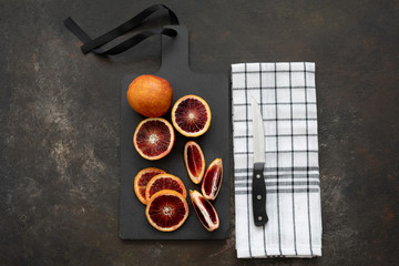 Top View of Blood Oranges on a Black Cutting Board on Black Textured Background; Black and White Kitchen Towel with Knife beside Cutting Board