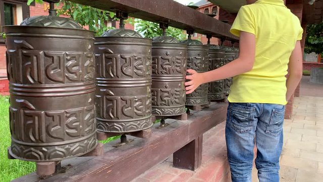 Caucasian boy touching, rotating nepalese traditional metal prayer wheels with mantra Om Mani Padme Hum, that means O, the jewel in the lotus, hanging in a row