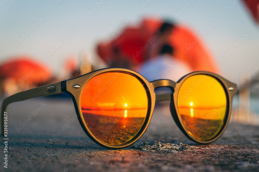 Wall mural Pair of sunglasses on the beach with the reflection of the sunset