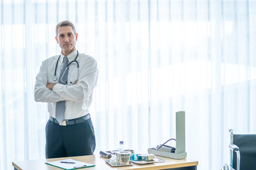 Smiling gray hair mature male doctor standing in hospital examination room with looking at camera. Confident professional general practitioner ready to work and consulting diagnosis medical healthcare